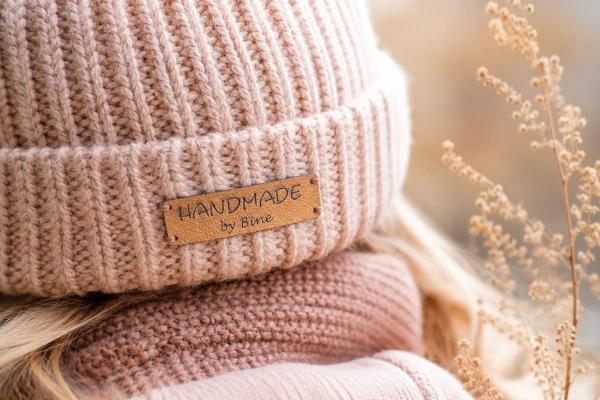 Rektangulär ljusbrun etikett i konstläder med "Handmade by Bine" på rosa stickad mössa.